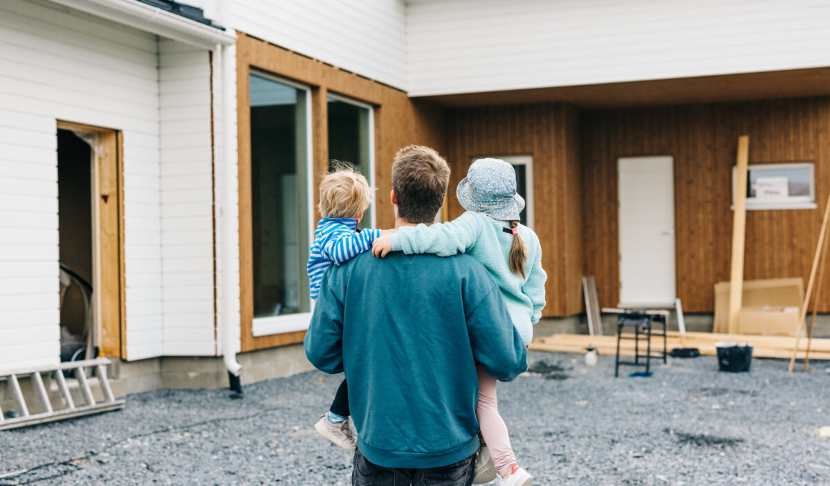 Mies pitelee lapsiaan sylissään, ja he katsovat rakenteilla olevaa omakotitaloa.