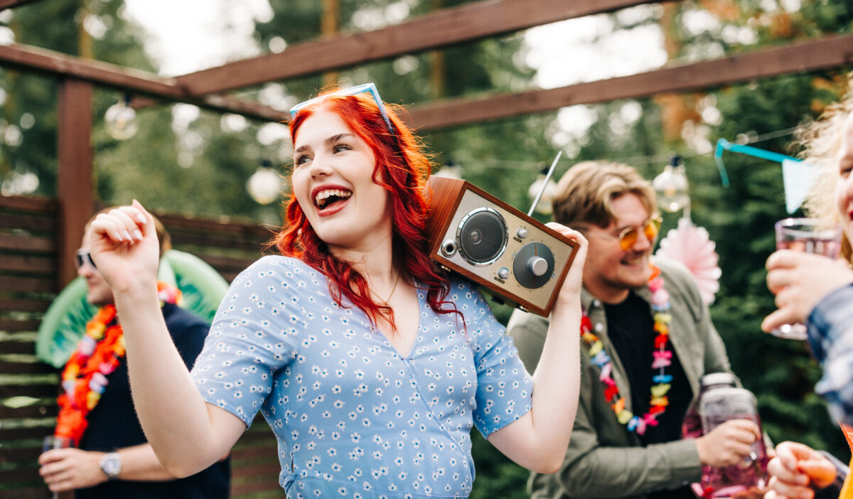 Nuori nainen radio olallaan kesäisessä juhlatunnelmassa, taustalla muitakin juhlijoita.